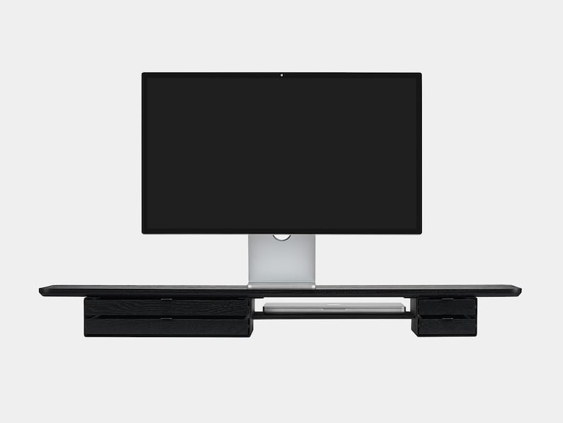 May include: A black computer monitor sits atop a black desk with two drawers on each side. The monitor has a silver stand. A laptop is visible on the desk between the drawers. The desk is mounted on a white wall.
