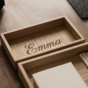 Può includere: Organizzatore da scrivania in legno con il nome "Emma" inciso in corsivo. L'organizzatore è realizzato in legno marrone chiaro e ha una forma rettangolare. Ulteriori scomparti contengono piccoli oggetti.