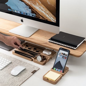 K&ouml;nnte beinhalten: Ein h&ouml;lzerner Schreibtisch-Organizer mit mehreren F&auml;chern f&uuml;r Stifte, Bleistifte, eine Uhr und ein Telefon. Der Organizer steht auf einem Holztisch mit einem Computermonitor, einem Notizbuch und einer Maus.