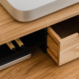 May include: Close-up of a wooden desk with a drawer and a black handle. The desk has a light wood grain finish. A white electronic device sits on top. Two pens are visible in a lower compartment.