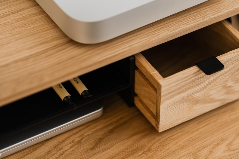 May include: Close-up of a wooden desk with a drawer and a black handle. The desk has a light wood grain finish. A white electronic device sits on top. Two pens are visible in a lower compartment.