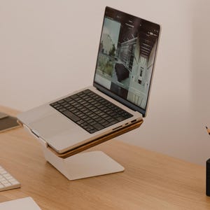 May include: A silver laptop computer is positioned on a white and wood laptop stand. The laptop screen displays a website with multiple images. A white wireless keyboard is placed in front of the stand, and a black pen holder sits to the right.