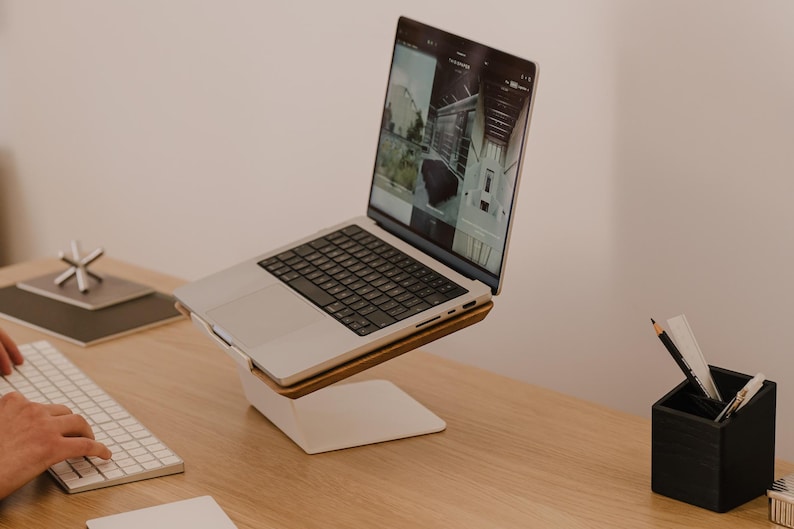 May include: A silver laptop computer is positioned on a white and wood laptop stand. The laptop screen displays a website with multiple images. A white wireless keyboard is placed in front of the stand, and a black pen holder sits to the right.