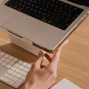 May include: A silver laptop computer resting on a white angled stand. The laptop features a black keyboard and a trackpad. A white wireless keyboard with square keys is positioned in front of the laptop stand on a light wood surface.
