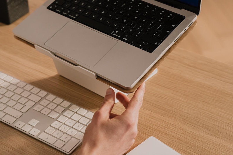 May include: A silver laptop computer resting on a white angled stand. The laptop features a black keyboard and a trackpad. A white wireless keyboard with square keys is positioned in front of the laptop stand on a light wood surface.