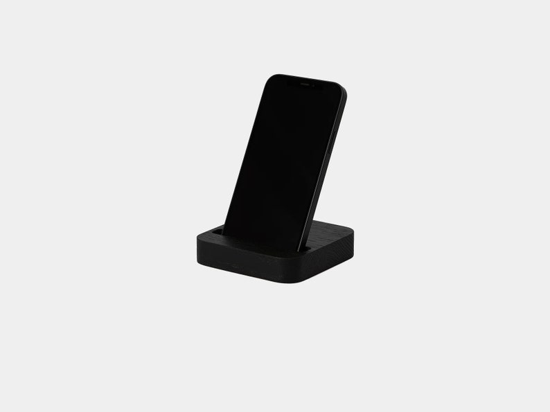K&ouml;nnte beinhalten: Ein schwarzer Smartphone-St&auml;nder aus Holz. Der St&auml;nder ist so konzipiert, dass er ein Telefon aufrecht h&auml;lt, mit einem Schlitz, in dem das Telefon ruhen kann.