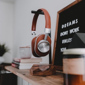 May include: Brown leather over-ear headphones with silver accents on a wooden stand. The headphones are resting on a wooden surface with a sign that reads "Dreams don't work unless you" in white lettering on a black background.