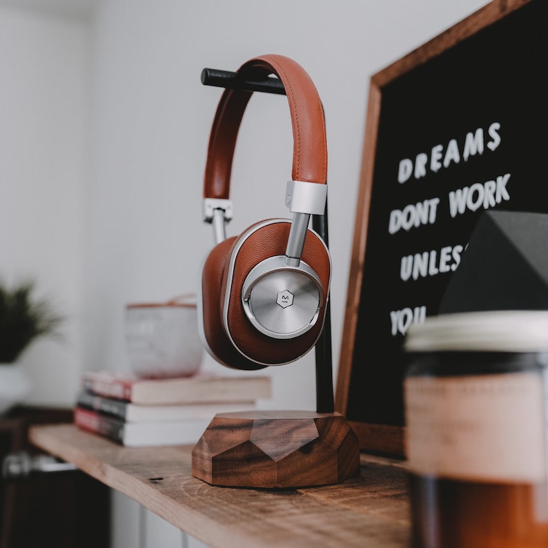 May include: Brown leather over-ear headphones with silver accents on a wooden stand. The headphones are resting on a wooden surface with a sign that reads "Dreams don't work unless you" in white lettering on a black background.