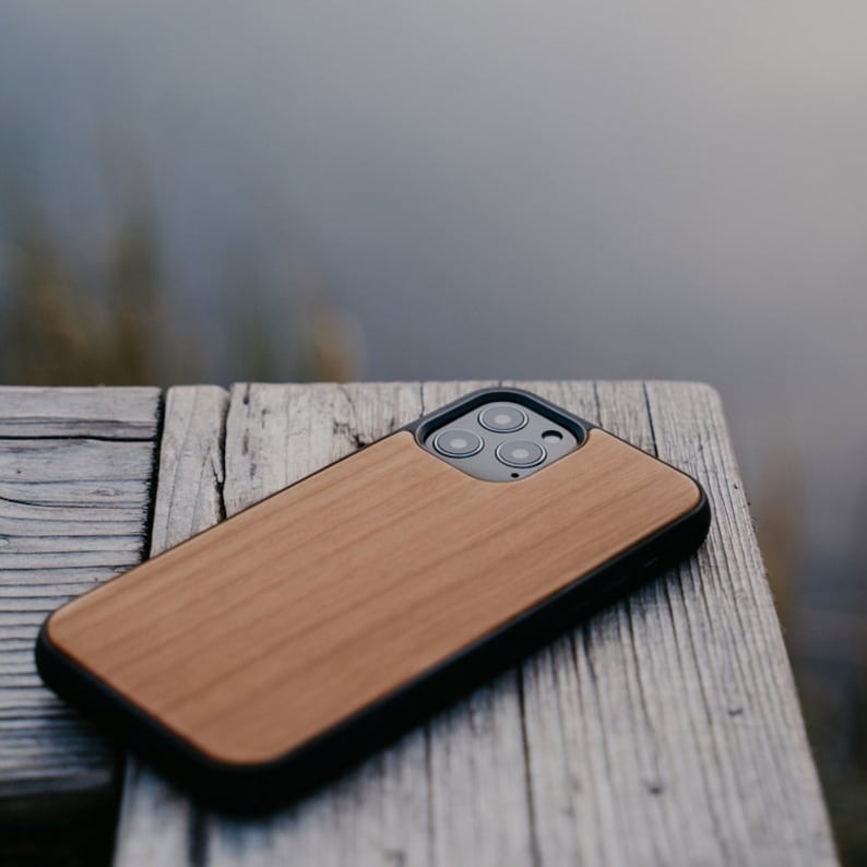 May include: A wooden phone case with a black trim and a camera cutout. The case is made of natural wood and has a light brown color.