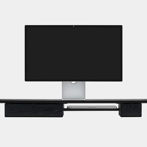 May include: A black wooden monitor stand with a built-in drawer and shelf, supporting a large computer monitor. The stand is designed to elevate the monitor and provide additional storage space. The monitor is silver and black.