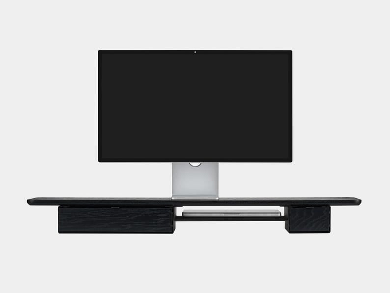 May include: A black wooden monitor stand with a built-in drawer and shelf, supporting a large computer monitor. The stand is designed to elevate the monitor and provide additional storage space. The monitor is silver and black.