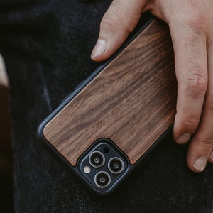 May include: A wooden phone case with a black trim and a camera lens cutout. The case has a natural wood grain pattern.