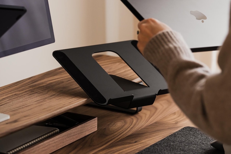 May include: A black metal laptop stand with a rectangular cutout, designed to elevate a laptop. The stand is on a wooden desk, with a monitor and a small wooden tray visible in the background. The stand is angled for ergonomic use.