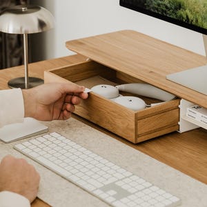 May include: A wooden desk with a built-in drawer holding white headphones. A silver desk lamp and a computer monitor are also visible. A white keyboard and mouse are on the desk.