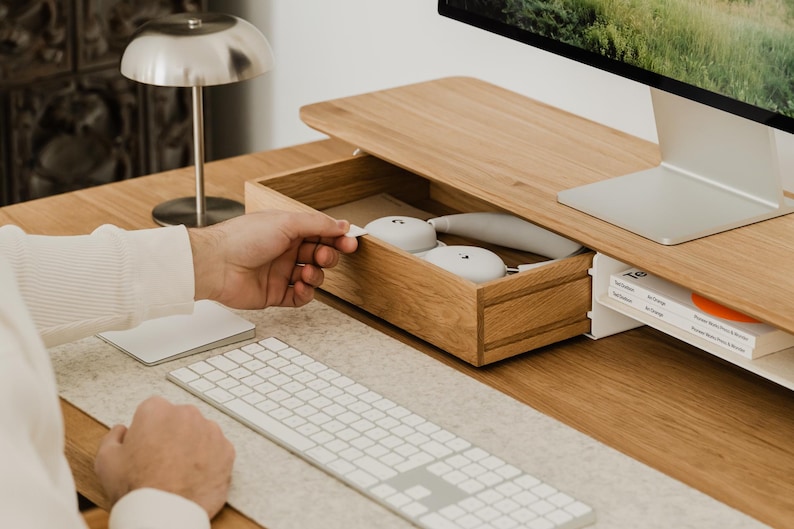 May include: A wooden desk with a built-in drawer holding white headphones. A silver desk lamp and a computer monitor are also visible. A white keyboard and mouse are on the desk.