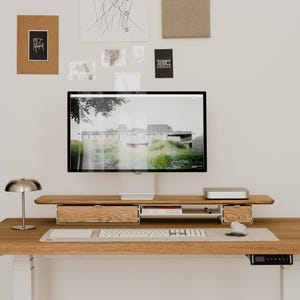 May include: A modern wooden desk with a computer monitor, keyboard, and mouse. The desk has two drawers and a small shelf. A silver lamp sits on the desk, and artwork hangs on the wall above.