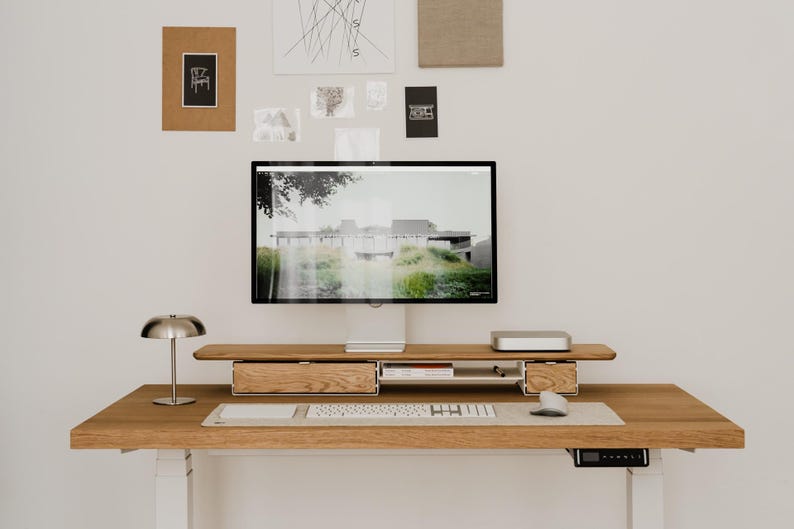 May include: A modern wooden desk with a computer monitor, keyboard, and mouse. The desk has two drawers and a small shelf. A silver lamp sits on the desk, and artwork hangs on the wall above.