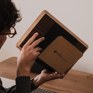May include: A rectangular tablet stand featuring a dark wood grain finish with layered plywood edges. The stand includes a square cork-textured back panel with a small black logo printed in the center, designed to support a tablet device securely.