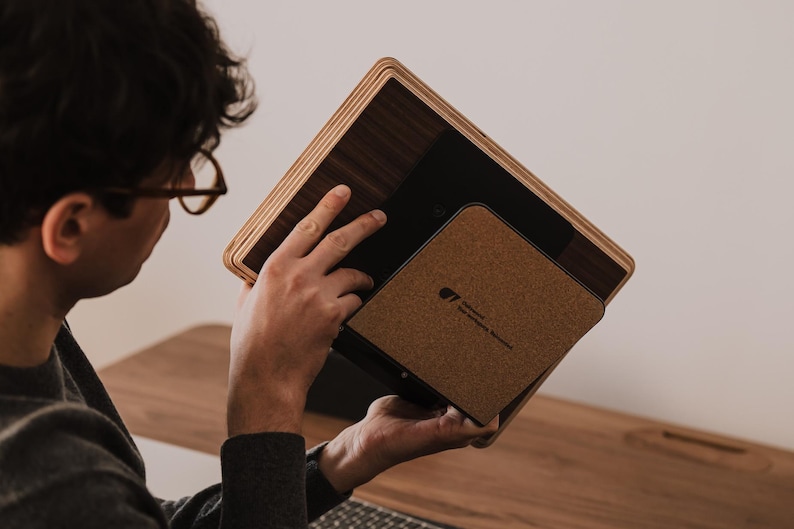 May include: A rectangular tablet stand featuring a dark wood grain finish with layered plywood edges. The stand includes a square cork-textured back panel with a small black logo printed in the center, designed to support a tablet device securely.