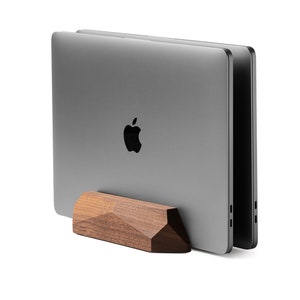 May include: A wooden laptop stand with a geometric design, holding two closed silver laptops. The stand is made of dark brown wood.