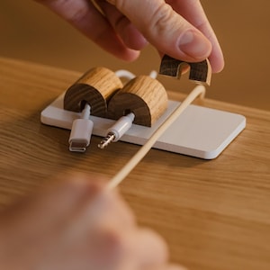 May include: Wooden cable tidy with two cylindrical holders and a white base. The tidy features a slot for each cable and a separate piece to secure the cables. The cables are white and the tidy is on a wooden surface.