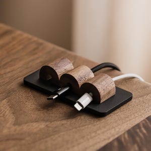 May include: A cable tidy with three wooden sections, each holding a charging cable. The organiser is made of dark wood and sits on a black base. The cables are black and white, and the tidy is on a wooden surface.