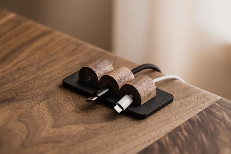 May include: A cable tidy with three wooden sections, each holding a charging cable. The organiser is made of dark wood and sits on a black base. The cables are black and white, and the tidy is on a wooden surface.