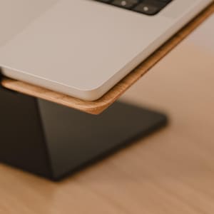 May include: A laptop stand featuring a matte black metal base and a light-toned wooden platform. The silver laptop rests securely on the wooden surface, which is angled upward to provide an elevated viewing position for the computer screen.