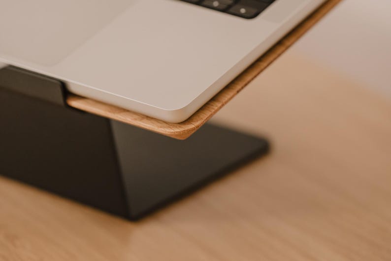 May include: A laptop stand featuring a matte black metal base and a light-toned wooden platform. The silver laptop rests securely on the wooden surface, which is angled upward to provide an elevated viewing position for the computer screen.