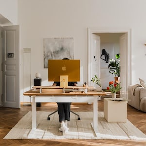 Op de afbeelding: Een moderne thuiskantooropstelling met een houten bureau en een grote gouden computermonitor. De kamer heeft een neutraal kleurenpalet, met een beige vloerkleed, witte muren en een lichtgekleurde bank. Decoratieve planten en kunstwerken voegen visuele interesse toe.