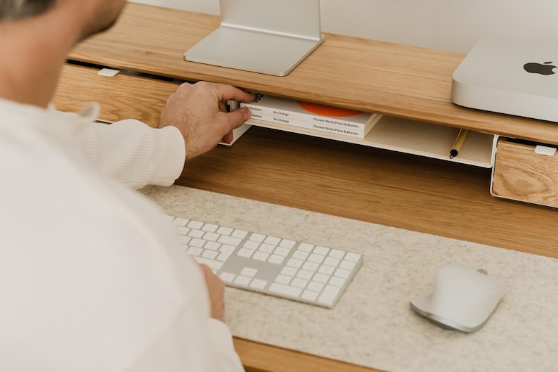 May include: A light wood desk with a built-in shelf holding a book with orange accents. A white keyboard, mouse, and computer monitor are on the desk. A person's hand is reaching for the book.