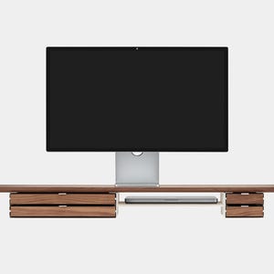May include: A modern wooden desk with integrated drawers and a shelf for a laptop. A large computer monitor is centered above the desk. The desk is made of dark wood and has a minimalist design.