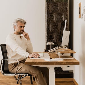 May include: A man sits at a wooden desk with a laptop, keyboard, and notebook. The desk is height-adjustable and has a white base. The man is wearing a cream-colored shirt and brown pants. A black chair is behind him.