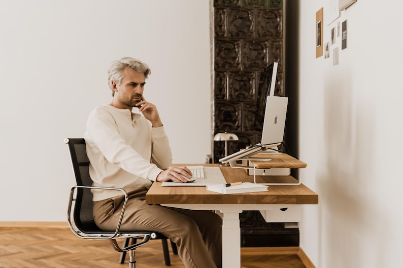 May include: A man sits at a wooden desk with a laptop, keyboard, and notebook. The desk is height-adjustable and has a white base. The man is wearing a cream-colored shirt and brown pants. A black chair is behind him.