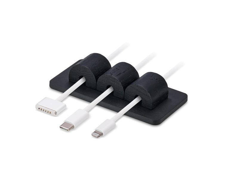 May include: A black cable tidy with three slots, holding white charging cables with different connector types. The tidy is rectangular with rounded edges, and the cable holders are cylindrical.