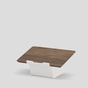 May include: A laptop stand featuring a rectangular dark brown wood grain platform supported by a solid white metal base. The stand has a tilted surface designed to hold a device at an angle, with a small white lip at the front edge to secure it.