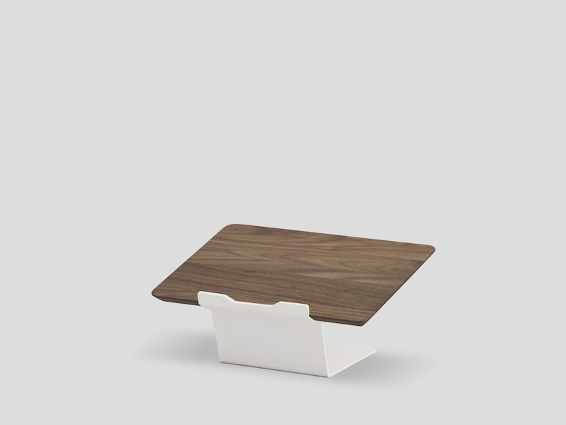 May include: A laptop stand featuring a rectangular dark brown wood grain platform supported by a solid white metal base. The stand has a tilted surface designed to hold a device at an angle, with a small white lip at the front edge to secure it.