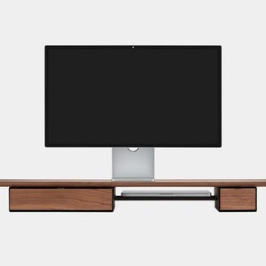 May include: A modern wooden monitor stand with two drawers and a shelf for a laptop. The stand is made of dark wood and has a sleek, minimalist design. A large black computer monitor sits on top of the stand.