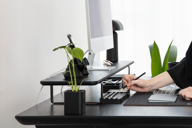 May include: A black wooden monitor stand with a metal frame, a black charging station with a phone and watch, a small potted plant, and a black pencil holder with pencils. The stand is on a black desk with a gray mousepad and a laptop.