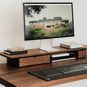 May include: A modern wooden desk setup featuring a computer monitor displaying a landscape image. The desk includes a keyboard, mouse, and a wooden monitor stand with storage. The desk is made of dark wood.