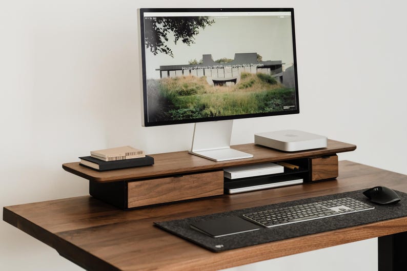 May include: A modern wooden desk setup featuring a computer monitor displaying a landscape image. The desk includes a keyboard, mouse, and a wooden monitor stand with storage. The desk is made of dark wood.