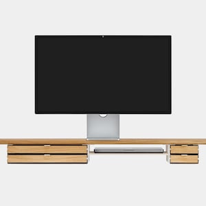 May include: A wooden computer monitor stand with a built-in shelf and drawers. The stand supports a large black screen monitor. The stand has two sets of drawers on either side and a shelf for a laptop.