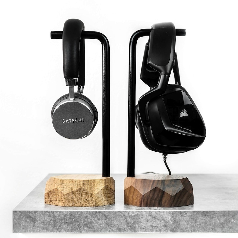 May include: Two black headphone stands with wooden bases. The stand on the left has a silver headphone with the word "SATECHI" on it. The stand on the right has a black headphone with a logo on it.