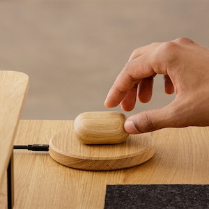 Pode incluir: Uma base de carregamento sem fio de madeira com uma caixa de madeira para fones de ouvido sem fio em cima. Uma mão está pairando sobre a caixa.