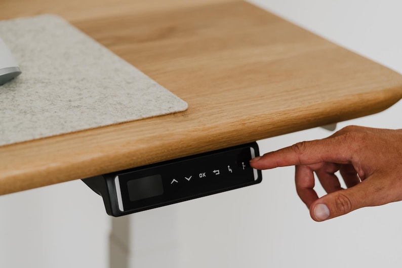 May include: A black control panel with a digital display and buttons for an adjustable standing desk. The buttons include up, down, OK, and memory presets.