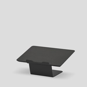 May include: A matte black metal laptop stand featuring a rectangular platform with a small front lip to secure a device. The stand has a bent, angled design that elevates the surface, providing a stable base for a laptop or tablet computer.