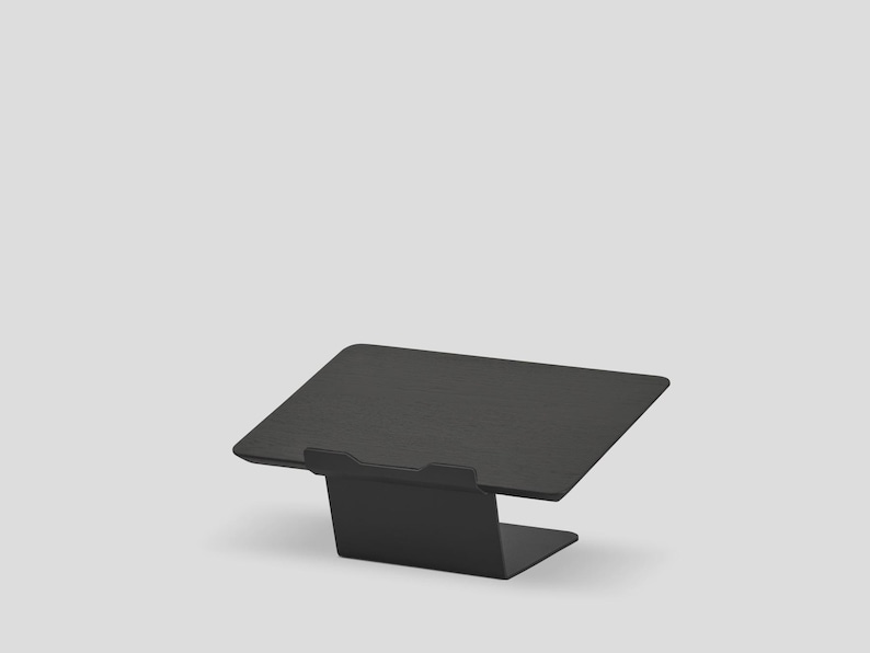 May include: A matte black metal laptop stand featuring a rectangular platform with a small front lip to secure a device. The stand has a bent, angled design that elevates the surface, providing a stable base for a laptop or tablet computer.
