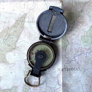 May include: A black folding sighting compass with a circular dial featuring green and black markings, a brass-coloured metal thumb loop, and a hinged lid with a vertical sighting wire, resting on a topographic map with contour lines and text.