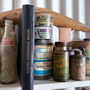 May include: A shelf displays a collection of vintage automotive products and a book titled "MOTOR ENGINEERING-CHARTS". The items include cans of wax, lacquer thinner, and motor oil, alongside a glass bottle. The items show signs of age and wear.