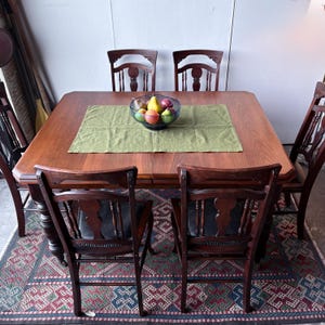 May include: A square wooden dining table with a smooth brown finish and four matching chairs featuring spindle backs and dark seats. A green rectangular fabric runner sits in the centre, topped with a glass bowl containing several colourful fruits.